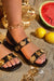 Black sandals with gold embellishments worn by a person, with a gold chain and watermelon in the background.